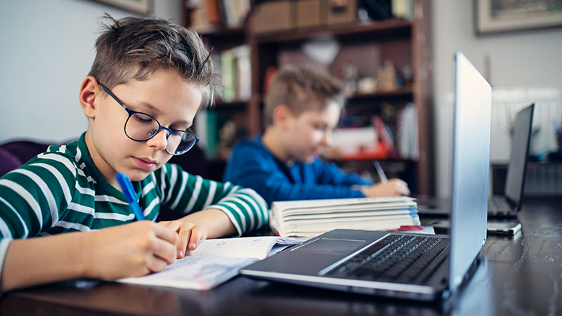 Dois rapazes sentados a uma mesa, em casa, escrevem num caderno e têm um computador aberto à sua frente
